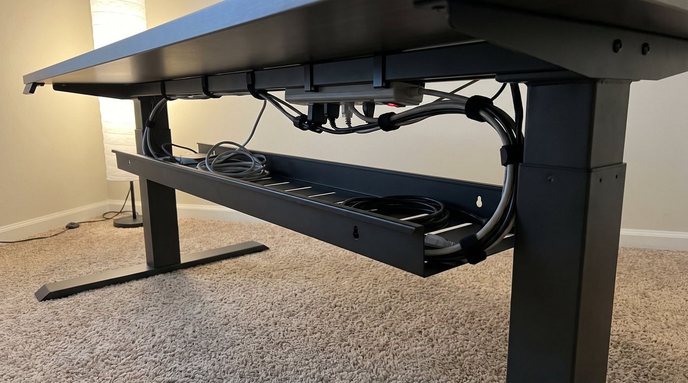 Underside view of a standing desk showing a metal cable tray, a mounted power strip, and cables routed with velcro ties toward one column
