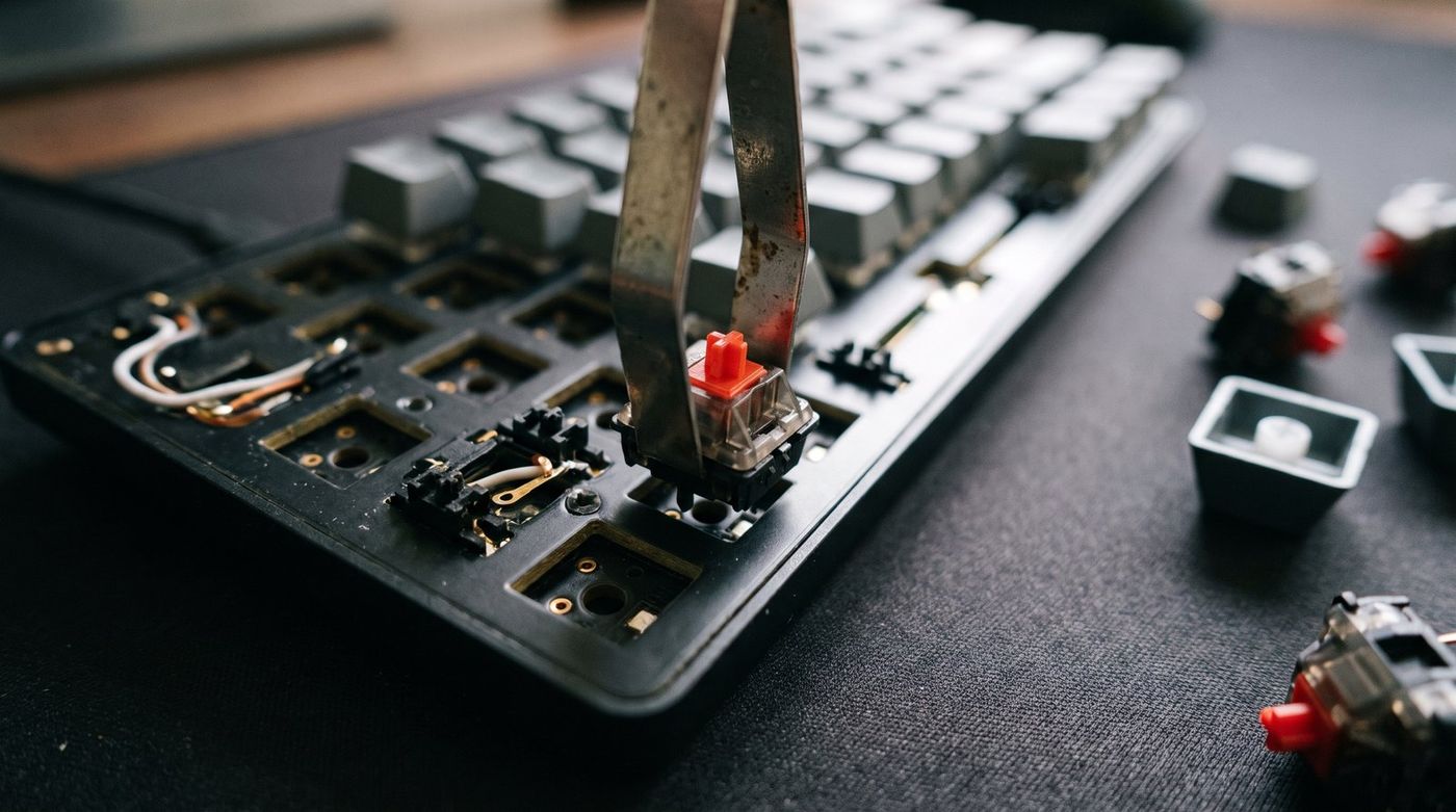 Close-up of a mechanical keyboard with a switch puller lifting one switch out of a hot-swap socket, keycaps scattered nearby