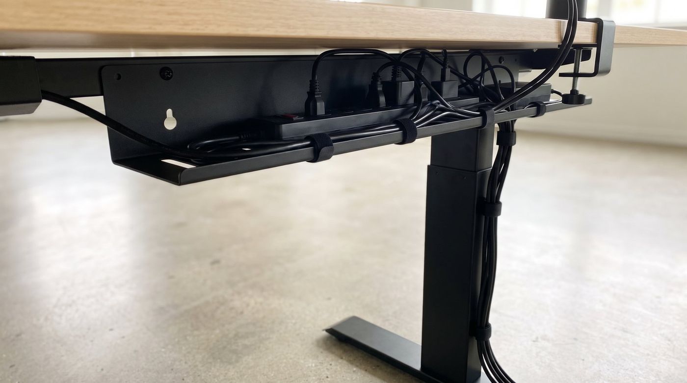 Underside of a standing desk showing a metal cable tray holding a power strip with cables bundled by velcro ties